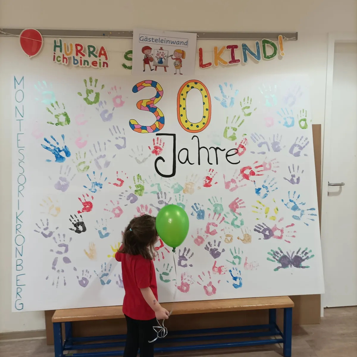 Ein Kind mit grünem Luftballon steht vor der Gästeleinwand: Hunderte bunte Handabdrücke rund um die Aufschrift „30 Jahre“, am Rand „Montessori Kronberg“.
